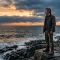 A rugged man standing at dawn on a rocky shoreline, composed and still — representing masculine identity and mindful strength