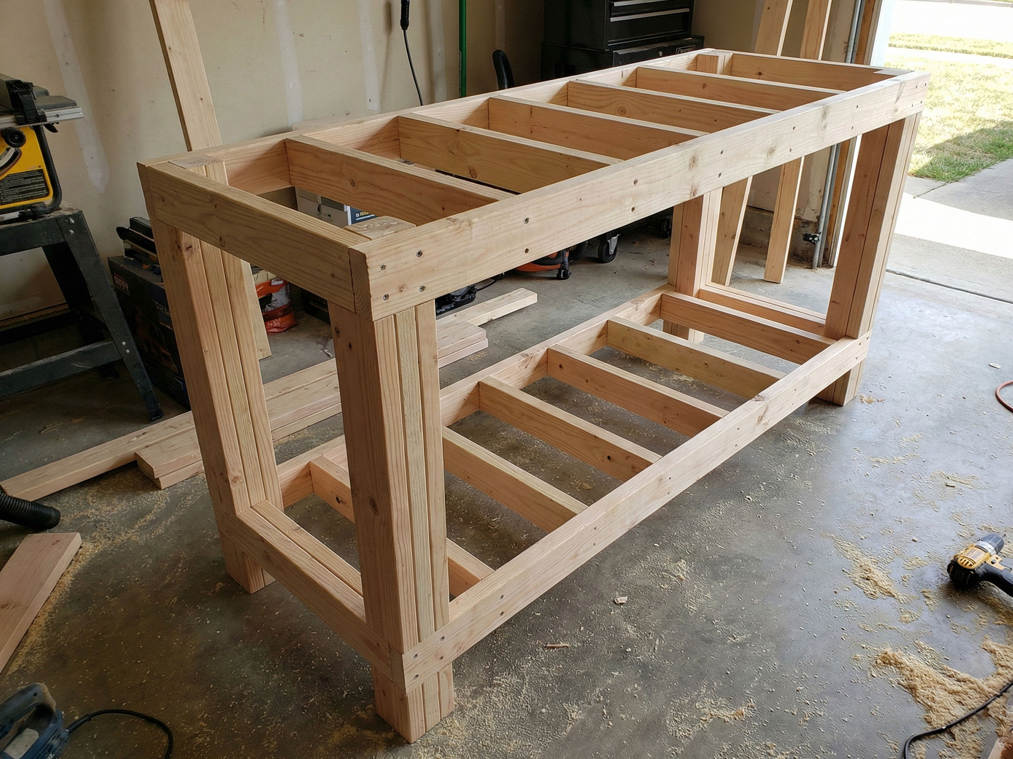 A sturdy workbench skeleton standing upright in a workshop showing massive laminated 2x4 legs and the lower shelf support framing.