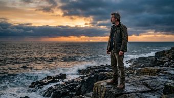 A rugged man standing at dawn on a rocky shoreline, composed and still — representing masculine identity and mindful strength