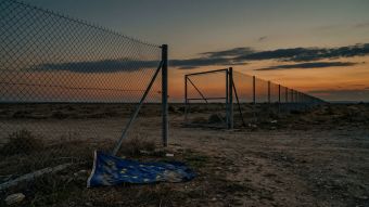 A wire border fence stretches across an open landscape at dusk, with the gate standing open and no enforcement presence visible — a visual metaphor for Europe's immigration enforcement gap.