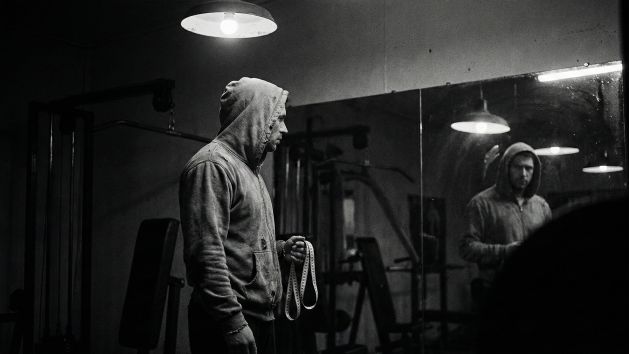 Un hombre examinando su reflejo, simbolizando la presi&oacute;n de las estad&iacute;sticas f&iacute;sicas masculinas.